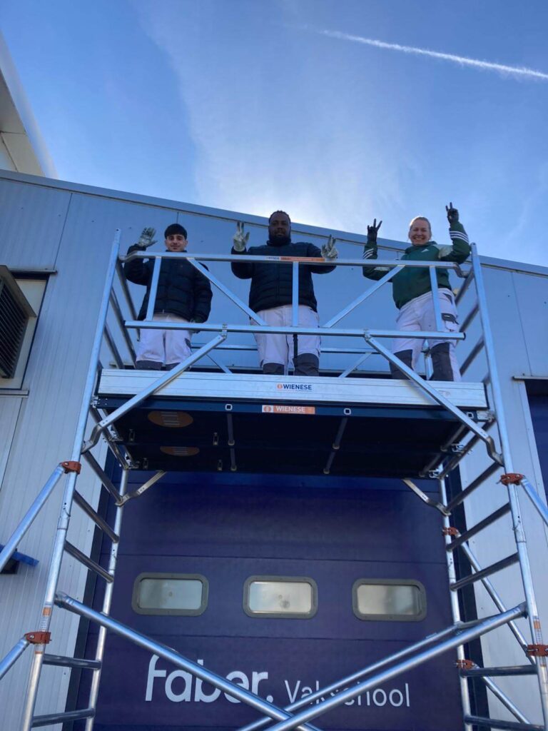 Drie leerling-schilders op een zelfgebouwde steiger tijdens de praktijkles steigerbouwen.